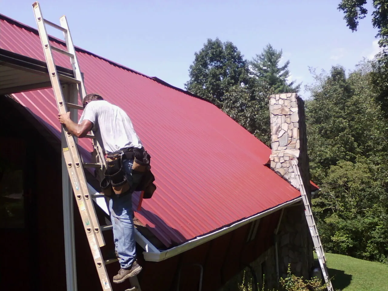 Roofer on a ladder beside a red metal roof and stone chimney — Soffit & Fascia Repair by Roofing Brothers in Conshohocken